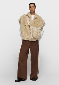 Chaleco de piel sintética beige con bolsillo con cremallera y correa a juego, combinado con pantalones de pierna ancha marrón oscuro con rayas laterales y zapatos negros.