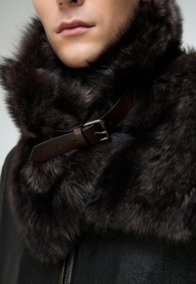 Tumepruun karvakaelus paksu tekstuuriga, kinnitatud pruuni nahast rihmaga, millel on hõbedane pandel, kinnitatud musta nahktagi külge.