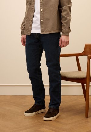 Giacca marrone a bottoni sopra una maglietta bianca, abbinata a jeans di denim scuri e sneakers marroni con suole bianche, in piedi accanto a una sedia di legno.