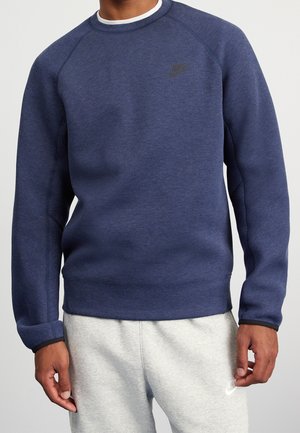 Mann trägt einen dunkelblauen Nike-Pullover und hellgraue Jogginghosen mit Nike-Logo, steht vor einem schlichten Hintergrund.