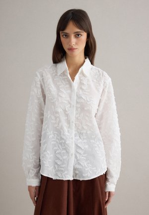 Junge Frau trägt eine weiße langärmelige bestickte Bluse mit Knöpfen und einen braunen Rock, steht vor einem einfarbigen beigen Hintergrund.