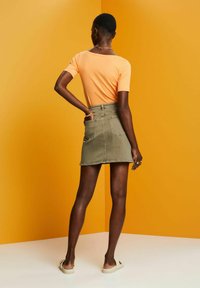 Oranges T-Shirt und olivegrüner Jeansrock mit hinteren Taschen, kombiniert mit hellen Sandalen vor einem gelben Hintergrund.