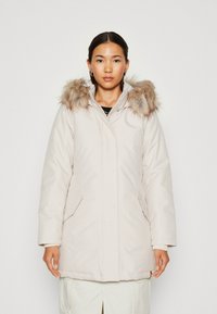 Parka beige clair avec capuche en fausse fourrure. Dotée d'une fermeture éclair, de poches latérales et d'un design légèrement rembourré pour plus de chaleur. Texture lisse et mate.