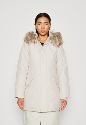 Parka beige chiaro con cappuccio in eco-pelliccia. Presenta una chiusura con zip, tasche laterali e un design leggermente imbottito per il calore. Texture liscia e opaca.