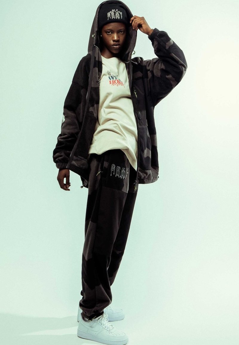 Sudadera con capucha de camuflaje negro y gris con cremallera, camiseta blanca, pantalones de chándal negros con logo y zapatillas blancas. Tejido suave con un corte relajado.