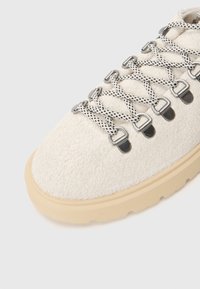 Semišová obuv v off-white farbe s textúrovaným povrchom, zdobená čiernymi a bielymi šnúrkami a kovovými okenkami, doplnená béžovou gumovou podrážkou.