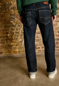 Jeans din denim albastru închis cu croială dreaptă, având buzunare spate și cusături deschise. După glezne cu o margine răsucită, cu adidași albi vizibili.