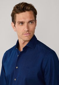 Camicia blu scuro con bottoni in tessuto morbido, con colletto classico, dettaglio discreto del logo sul lato sinistro del petto e texture liscia.