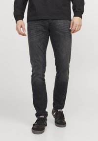 Jeans oscuros en corte slim con textura desgastada, combinados con zapatillas negras con parte superior de tela y suela de goma, mostrando un estilo casual.