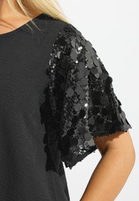 T-shirt nera con maniche corte impreziosite da paillettes nere lucide, creando un aspetto texturizzato e arricchito. La base in tessuto liscio contrasta con le paillettes.