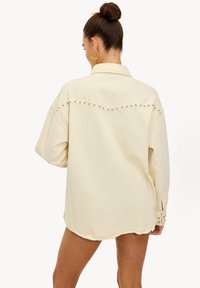 Camisa sobre tamaño de color crema con cuello, detalles de tachuelas a lo largo de la costura trasera y puños con botones, hecha de un tejido suave.