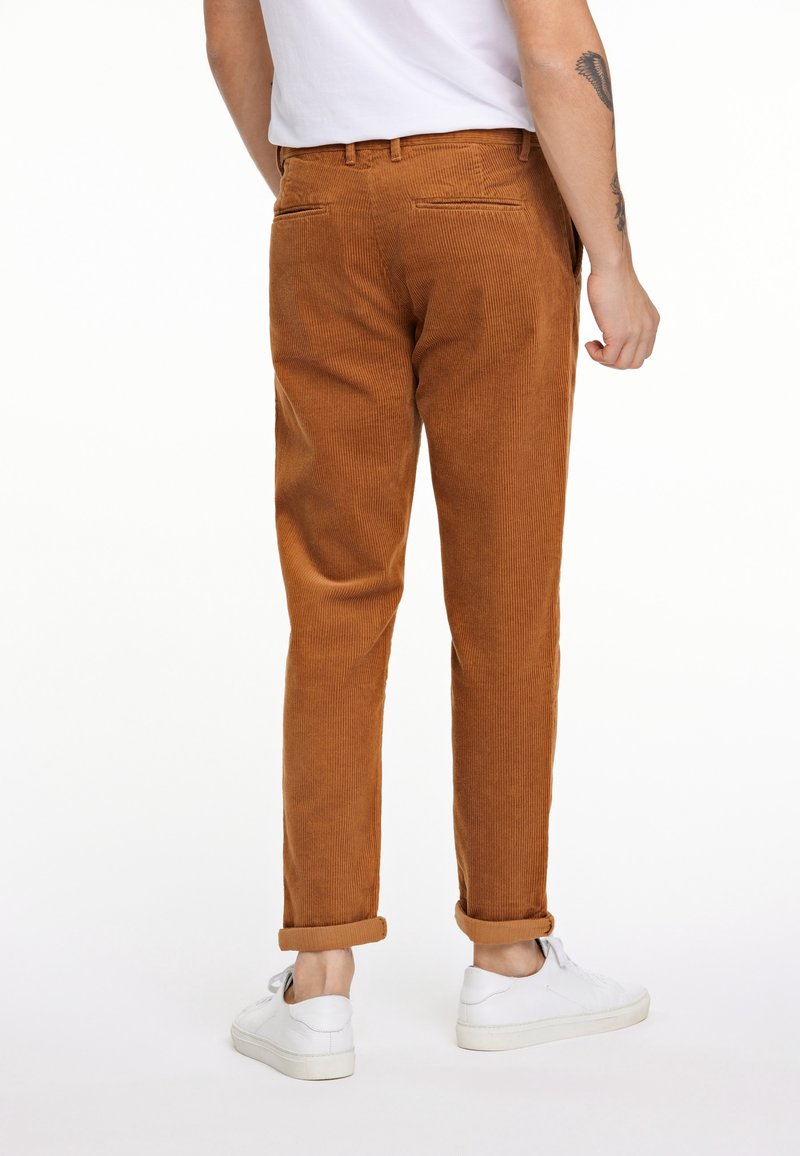 Homme portant un pantalon en velours côtelé marron retroussé et des baskets blanches, debout dos à la caméra sur un fond blanc.