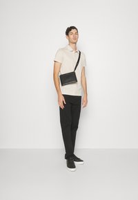 Bolso cruzado negro con textura acanalada vertical, correa ajustable, llevado sobre un polo beige claro y pantalones negros, con un fondo blanco.
