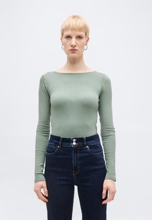 Femme aux cheveux blonds courts portant un haut à manches longues côtelé vert clair et un jean taille haute bleu foncé, se tenant devant un fond blanc.