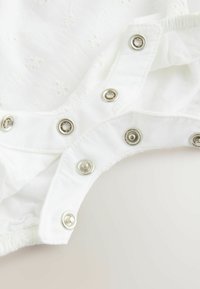 Vêtement en tissu blanc présentant une broderie florale et des boutons-pression argentés le long des ouvertures des jambes, avec des bords élastiqués froncés.