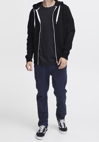 Felpa con cappuccio nera con zip e coulisse bianche, abbinata a una t-shirt nera, pantaloni blu navy e sneakers nere. Vestibilità casual e rilassata.