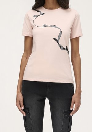 Kobieta ubrana w jasnoróżowy t-shirt z krótkim rękawem z czarnym wzorem wstążki oraz czarne spodnie typu cargo z bocznymi kieszeniami.