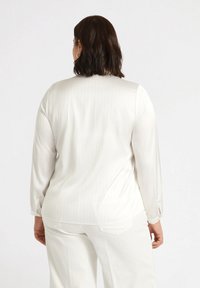 Blusa bianca a maniche lunghe con righe verticali, texture liscia e polsini a spacco, abbinata a pantaloni bianchi. Vista posteriore.