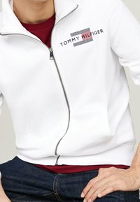 Sudadera blanca con cremallera hecha de tela suave, que presenta un logo a rayas rojas y azules en el pecho. Tiene cremallera en la parte delantera y bolsillos laterales.