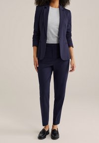 Námořnický modrý oblekový sako s knoflíkem, spárované s odpovídajícími kalhotami na míru. Šedé tričko s kulatým výstřihem pod ním. Černé polobotky dokončují vzhled.