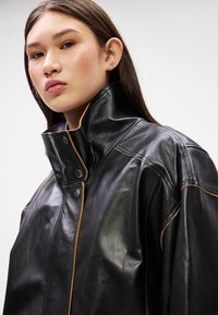 Crna kožna jakna s visokim ovratnikom, koja ima kontrastne narančaste šavove i kopčanje na dugmad. Glatka tekstura i oversized kroj.
