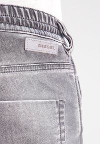 Szare dżinsy z elastycznym paskiem w talii, z białą skórzaną łatą z logo "DIESEL" oraz detalem kieszeni z tyłu.