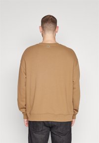 Hombre de espaldas vistiendo una sudadera color camel con un pequeño logo de cocodrilo verde en la parte superior de la espalda y jeans oscuros.