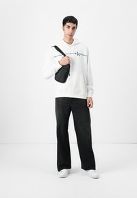 Witte hoodie met een logodetail, zwarte schoudertas, zwarte jeans en witte sneakers, staand tegen een plain witte achtergrond.