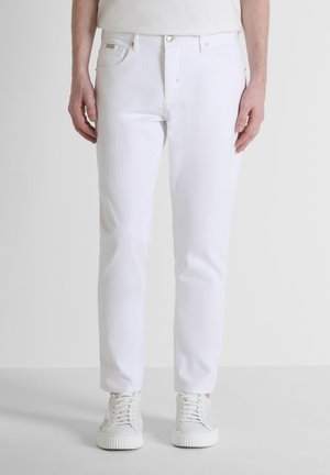 Man draagt slanke witte spijkerbroek en witte veter sneakers en staat tegen een effen achtergrond.