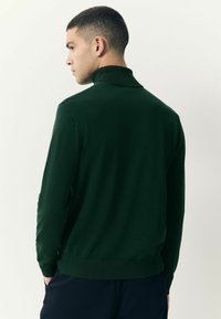 Pulover verde închis cu guler înalt, mâneci lungi și manșete răsucite. Textură netedă, formă ajustată și design de culoare uniformă.