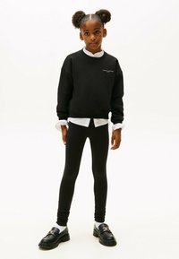 Niña joven con cabello voluminoso, vistiendo un sudadera negra, una camisa blanca con cuello, leggings negros y mocasines negros con calcetas blancas, de pie.