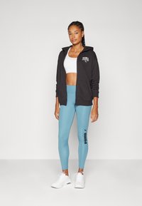 Sudadera negra con cremallera y logo, top corto blanco y leggings azul claro con un logo en el lateral. Zapatillas deportivas blancas completan el conjunto.