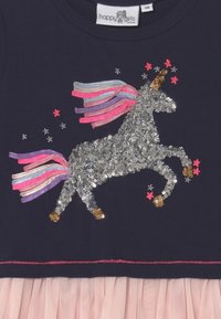 Paljettädd enhörning med flerfärgad man och gyllene hovar på marinblått tyg, ihopparad med rosa tyllkjol nedanför. Etiketten säger "glada flickor".