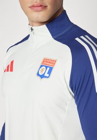 Bijela i tamnoplava sportska jakna na zatvaranje s patentnim zatvaračem, s logom Olympique Lyonnais i tri bijele pruge na tamnoplavim rukavima.