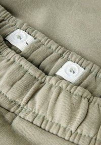 Zbliżenie na regulowany elastyczny pas w jasnozielonych spodniach z białym guzikiem i elastycznym paskiem do dopasowania rozmiaru.