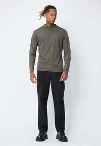 Un homme avec des dreadlocks porte une chemise à manches longues olive, un pantalon noir et des chaussures noires épaisses, se tenant devant un fond blanc.