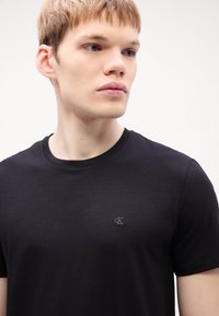 Black short-sleeve t-shirt made of soft cotton fabric, featuring a subtle embroidered logo on the left chest. Simple round neckline.