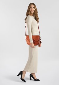 Borsa a tracolla in pelle marrone con pattina curvata, accessori decorativi e tracolla regolabile, indossata con un vestito beige a maniche lunghe e tacchi neri.