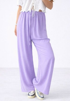 Personne portant un pantalon lavande ample, taille haute, avec cordon de serrage et des baskets blanches, associé à un top court blanc.