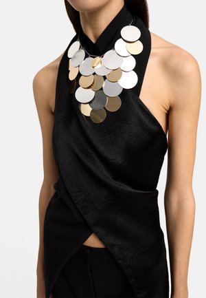Kobieta w czarnym, bezrękawnikowym topie na szyję z dużym naszyjnikiem wykonanym z nakładających się na siebie błyszczących złotych i srebrnych okrągłych dysków.