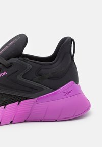 Zapatilla deportiva negra y morada con suela texturizada, parte superior de malla y collar acolchado en el talón, mostrando el logo de Reebok en el talón y el lateral.