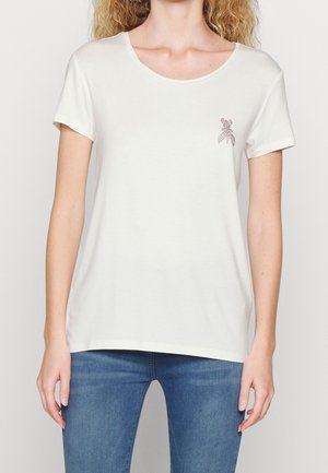 Žena v krátkém bílém tričku s kulatým výstřihem a malým dekorativním emblémem na levé straně hrudi, doplněná modrými džíny.