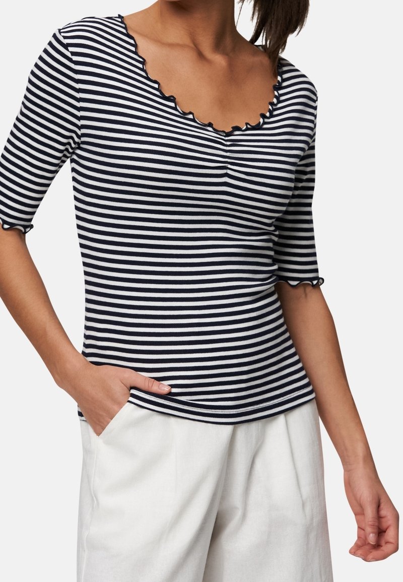Femme portant un haut à manches courtes rayé horizontal noir et blanc avec des bords festonnés et un pantalon blanc, main dans la poche.