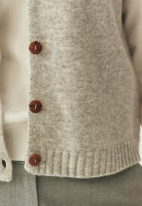 Gray knit cardigan with a ribbed hem and five brown buttons. Worn over a white top, showcasing soft texture and casual design.