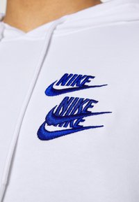Vit hoodie med blått broderat "NIKE"-logotyp tre gånger, med en smidig textur och klassisk design.