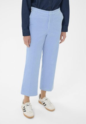 Person in einer hellblauen, weiten Cordhosen, einem dunkelblauen Langarmshirt und weißen Sneakern mit schwarzen Streifen vor weißem Hintergrund.
