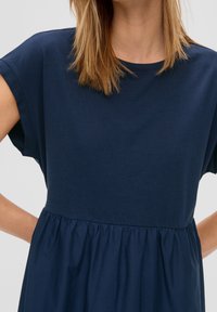 Tummansininen mekko, jossa on lyhyet hihat, muotoiltu yläosa ja poimutettu hame. Valmistettu pehmeästä kankaasta, jossa on sileä rakenne.