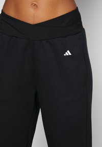 Záběr na střed těla osoby oblečené v černých teplácích Adidas s širokým pasem a bílým logem Adidas na levém bocím.