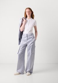 Wit poloshirt met drie knopen, lichtgrijze cargobroek met grote zakken en lichte sneakers. Model houdt een grijze jas vast.