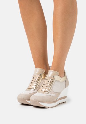 Jambes portant des baskets compensées beige avec des panneaux en tissu et dorés brillants, des semelles intermédiaires blanches et des semelles extérieures en caoutchouc beige sur un fond clair.